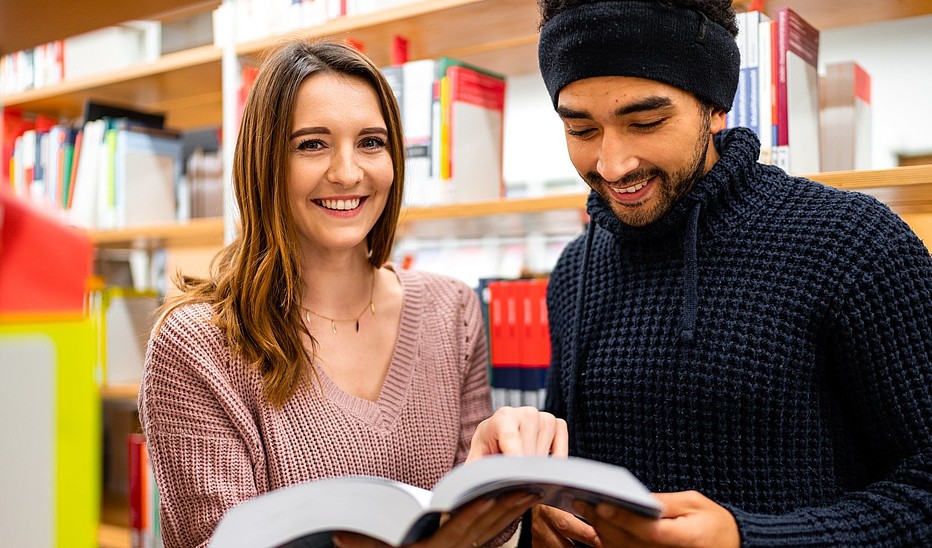 Zwei Studierende, eine junge Frau und ein junger Mann mit Migrationshintergrund stehen in der Bibliothek und schauen sich ein Buch an.