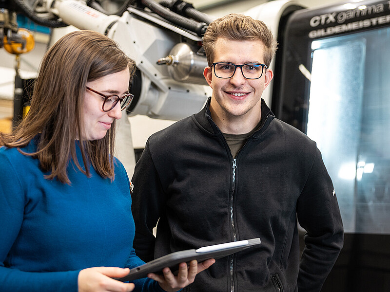 Zwei Studierende stehen an einer Anlage mit mechanischem Greifarm. Die Studentin schaut auf ein Tablet.