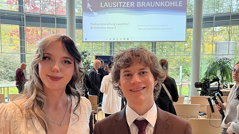 Zwei Studierende stehen in einem Veranstaltungssaal und lächeln in die Kamera.