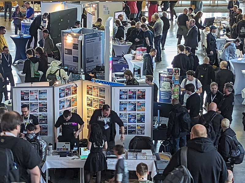 Viele Menschen in einem großen Raum auf einer Messe mit vielen Ständen, Plakaten und Aufstellern