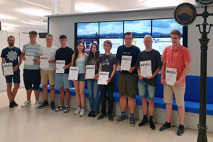 The students hold their certificates in their hands in the electrical engineering laboratory and look into the camera.