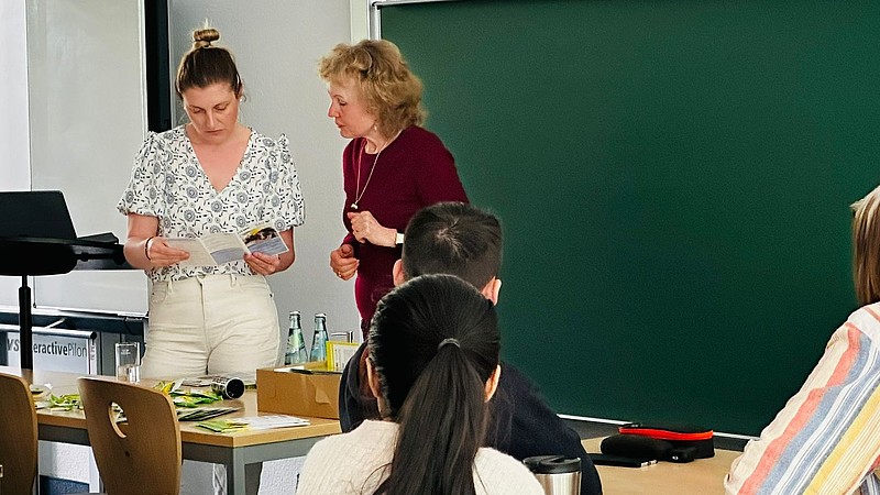 Kristin Groß und eine Studienkollegs-Mitarbeiterin aus Leipzig stehen im Seminarraum vor einer Tafel und sehen sich einen Flyer an.