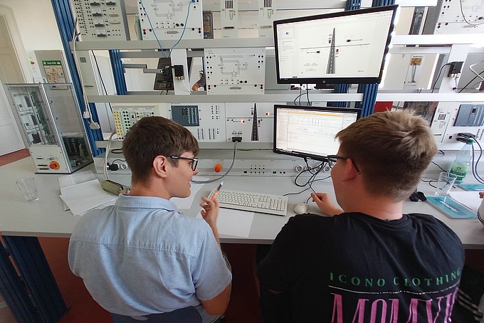 zwei junge Männer sitzen vor einem Versuchsstand mit mehreren Monitoren 