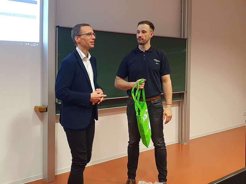 Prof. Kornhuber und Herr Schumann stehen vor der Tafel im Vorlesungssaal. Herr Schumann hält einen grünen Beutel in der Hand, während Prof. Kornhuber spricht.