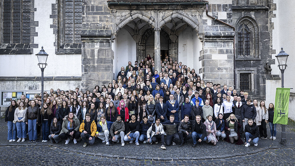 Foto: Philipp Herfort Eine große Menge junge Studierende und der Rektor stehen am Seiteneingang der Kirche auf den Stufen und dem vorderen Platz verteilt. Alle schauen in die Kamera.