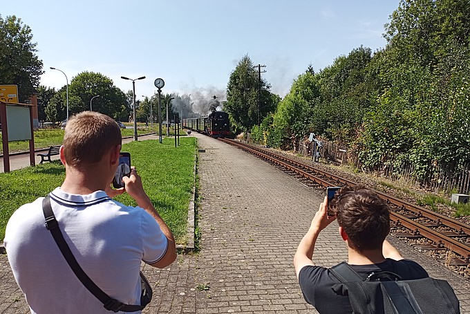 Zwei junge Männer stehen mit dem Rücken zur Kamera und fotografieren die einfahrende Bahn mit dem Handy