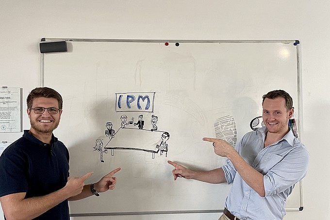 Jan-Hendrik und Karl zeigen vor einem Whiteboard auf die Aufschrift "IPM".