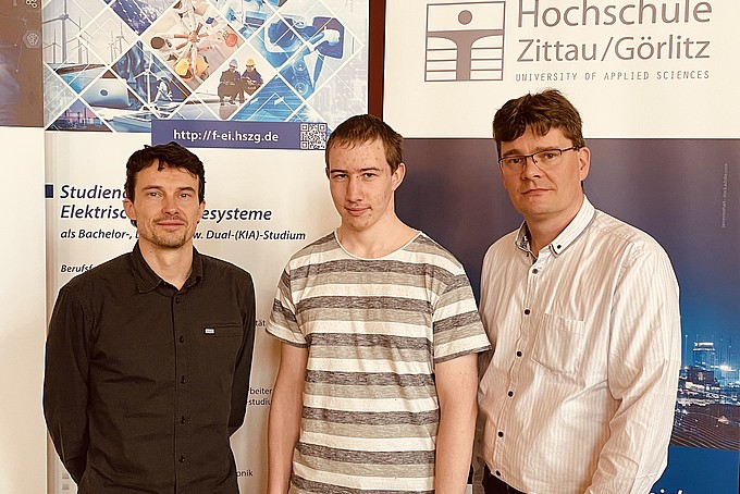 Drei Männer stehen vor einer Plakatwand mit der Aufschrift "Hochschule Zittau/Görlitz"